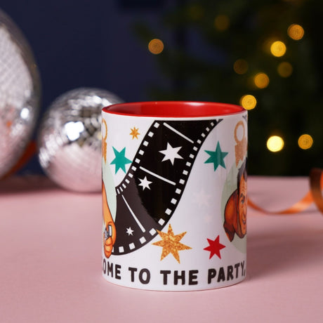 White 11oz gloss stoneware mug with red interior, film-strip and star print, cartoon character portraits, and "Welcome To The Party, Pal" text, shown near festive decorations.