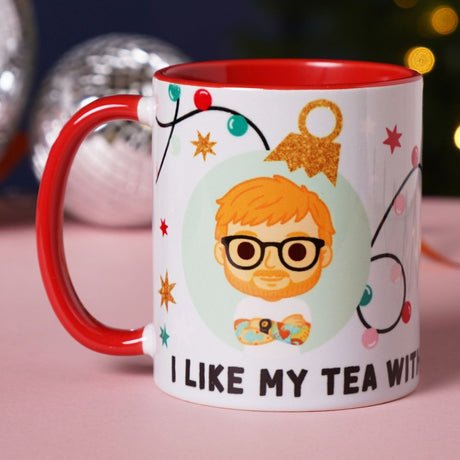 White stoneware mug with red handle and interior, featuring a cartoon ginger-haired bearded man in glasses amid Christmas lights and stars, with "I like my tea with..." text below.