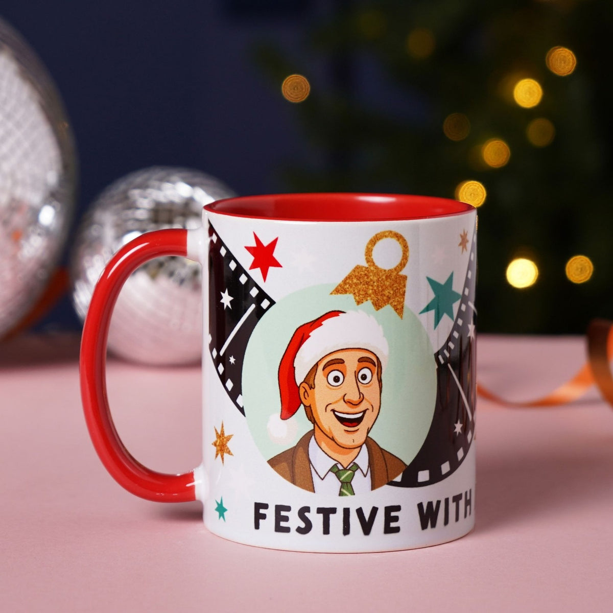 Stoneware mug with red interior and handle featuring a cartoon man in a Santa hat in a mint circle with film strips, stars, and Festive With text, shown beside baubles and lights.