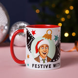 Stoneware mug with red interior and handle featuring a cartoon man in a Santa hat in a mint circle with film strips, stars, and Festive With text, shown beside baubles and lights.