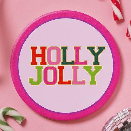 Round ceramic Christmas coaster with a bold pink rim and pale pink centre, featuring "HOLLY JOLLY" in multicoloured patch-style capitals with gold outlines, shown on a pink surface with candy canes.