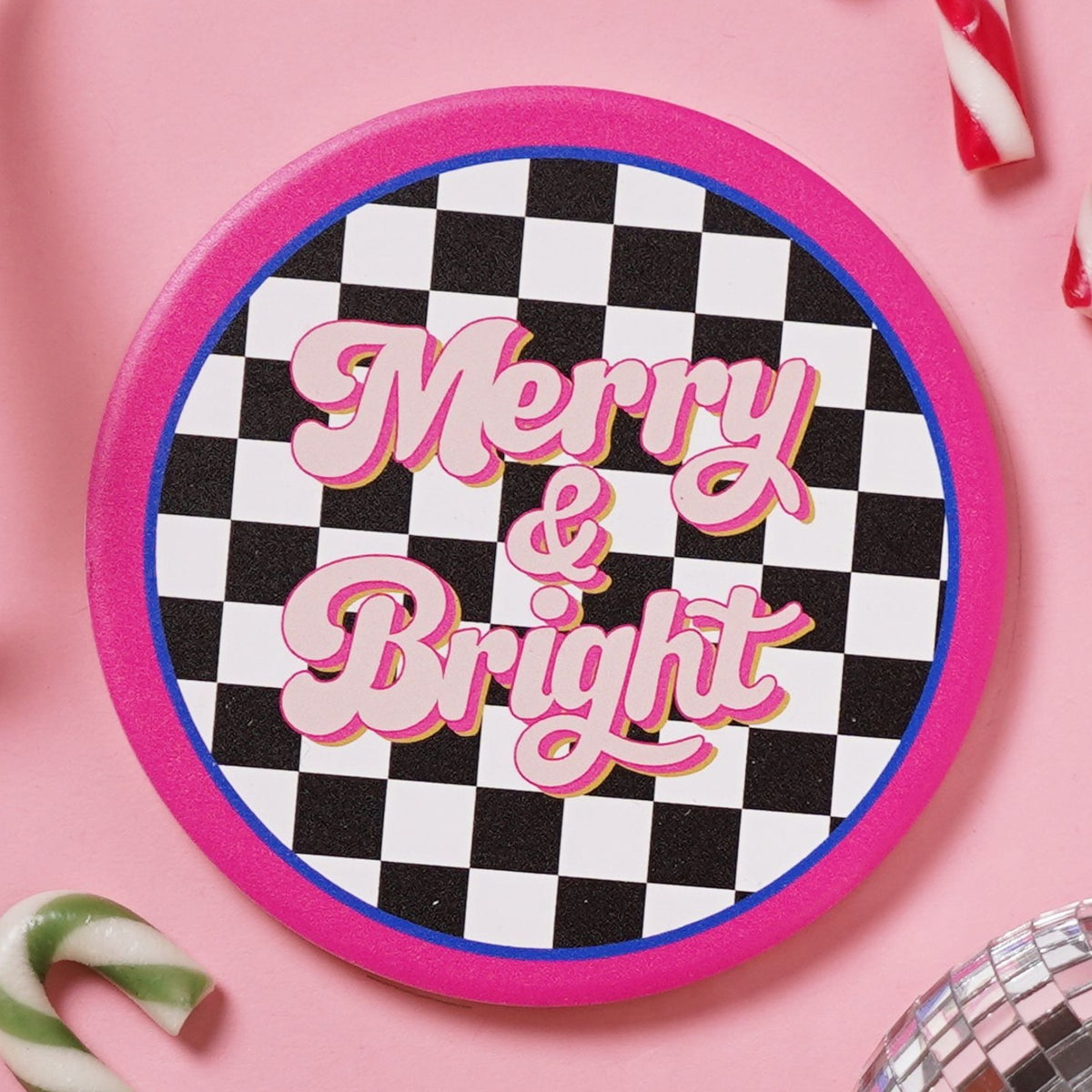 Round ceramic coaster with black and white chequerboard pattern, "Merry & Bright" in pink script, bordered by a hot pink rim and thin blue ring, on a pink background.
