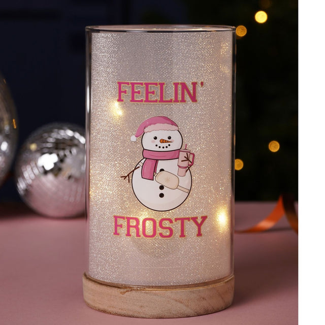 Light-up cylindrical decoration with white glitter finish, wood base, and pink-hatted snowman design printed with Feelin' Frosty in bold pink lettering, shown on a pink surface with bokeh lights.
