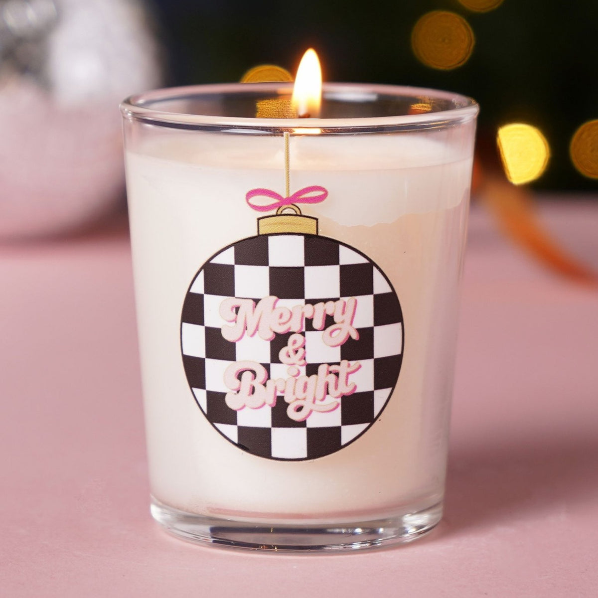 Merry & Bright 10cl votive candle in clear glass with white wax, lit wick, chequered bauble print, pink lettering, gold cap, and pink bow, set against a festive bokeh background.