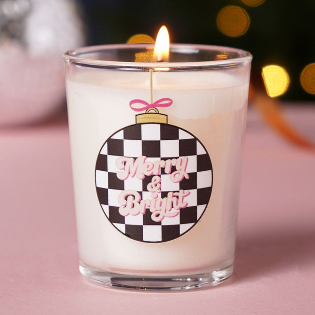 Merry & Bright 10cl votive candle in clear glass with white wax, lit wick, chequered bauble print, pink lettering, gold cap, and pink bow, set against a festive bokeh background.