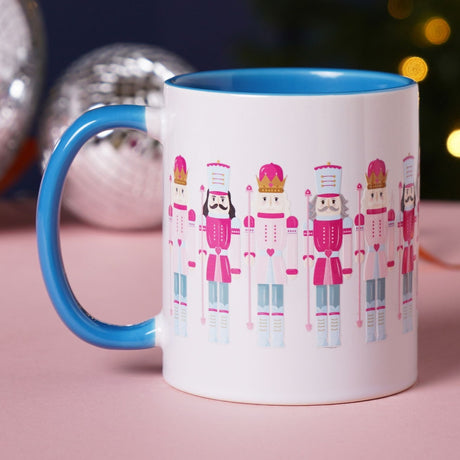 White stoneware mug with blue interior and handle, featuring a repeating pattern of pink and white nutcracker soldiers with gold crowns, shown beside festive baubles.