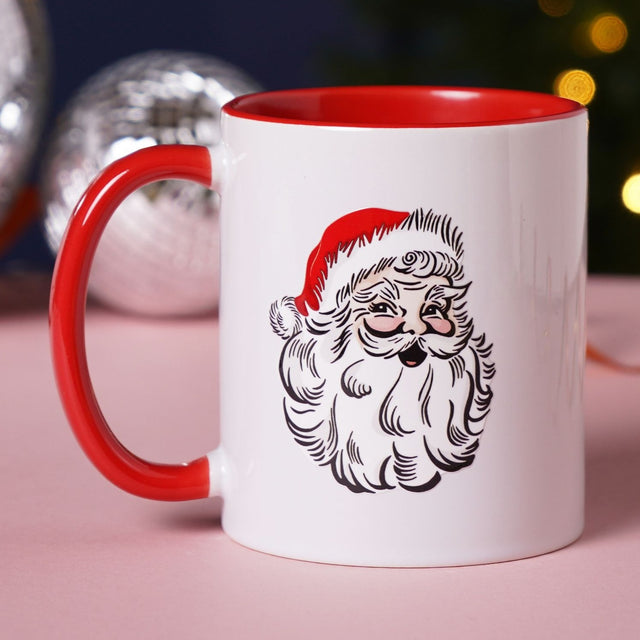 White stoneware 11oz mug with red handle and interior, featuring an illustrated Santa head with hat and beard, shown with Christmas baubles in the background.