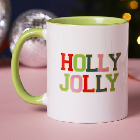 White stoneware mug with lime green handle and interior, featuring "HOLLY JOLLY" in bold multicoloured patch-style letters with gold outlines, 11oz, shown on a pink surface with festive decor.