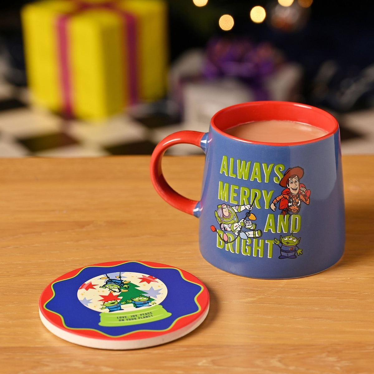 Toy Story Christmas porcelain mug in blue with red handle and interior, "Always Merry and Bright" text, Woody, Buzz and an alien, shown with matching round coaster on a wooden table.