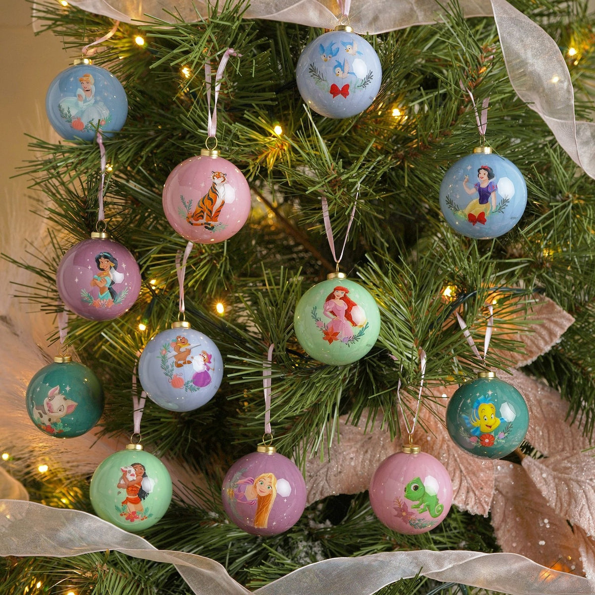 Set of 12 round glossy baubles in muted blue, pink and green tones, each printed with a Disney princess or sidekick design, shown hanging on a decorated Christmas tree.