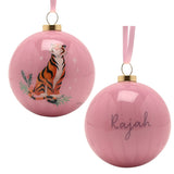Two glossy pink round baubles with gold caps and ribbon hangers, one showing Rajah the tiger with leaf accents, the other personalised with "Rajah" in script, on a white background.
