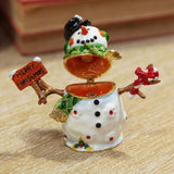 Christmas Snowman Metal Trinket Box with hinged lid open, white glossy finish, black top hat, green scarf, holding a Merry Christmas sign and candy cane, on a wooden surface.
