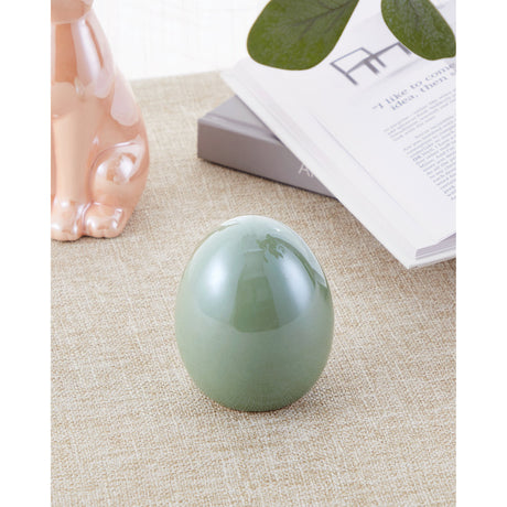 Yaypan ceramic decorative egg in glossy mint green, freestanding on a woven surface beside a rose gold rabbit figurine and an open book.