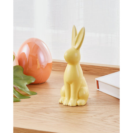 Yaypan ceramic rabbit ornament in glossy pastel yellow, sitting upright with tall ears, displayed on a wooden surface beside a glossy orange egg and a white book.