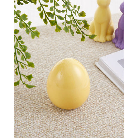 Yaypan Decorative Egg in glossy yellow, a smooth egg-shaped ornament on a flat base, shown on a woven tabletop beside a fern plant and small animal figurines.