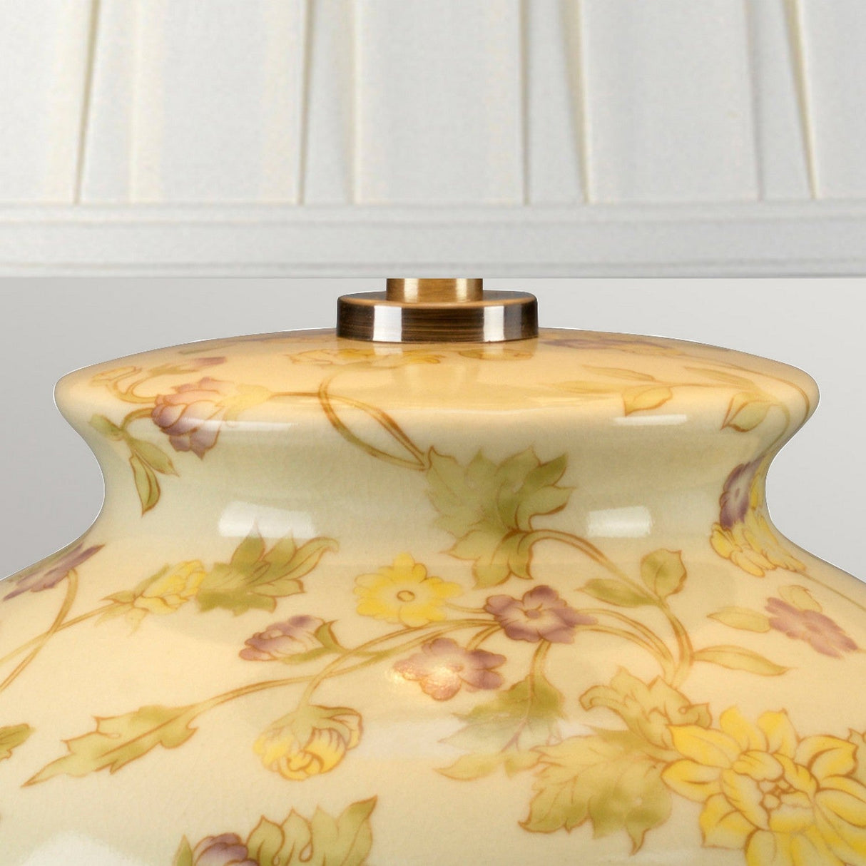 The Yellow Flowers 1 Light Table Lamp in yellow and purple features a close-up of its decorative Chinese porcelain base, adorned with a floral design showcasing yellow flowers and hues of green and purple. It is topped with a metallic fitting and paired with a pleated white lampshade in the background.