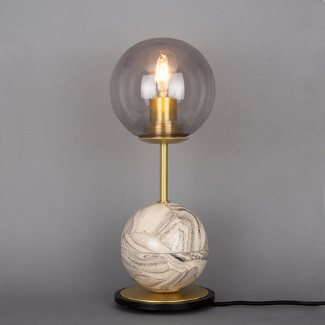 Zapp Ceramic Globe Table Lamp with smoked glass globe shade, brass stem, marbled grey and cream ceramic ball base on a matt black disc, set against a plain grey background.