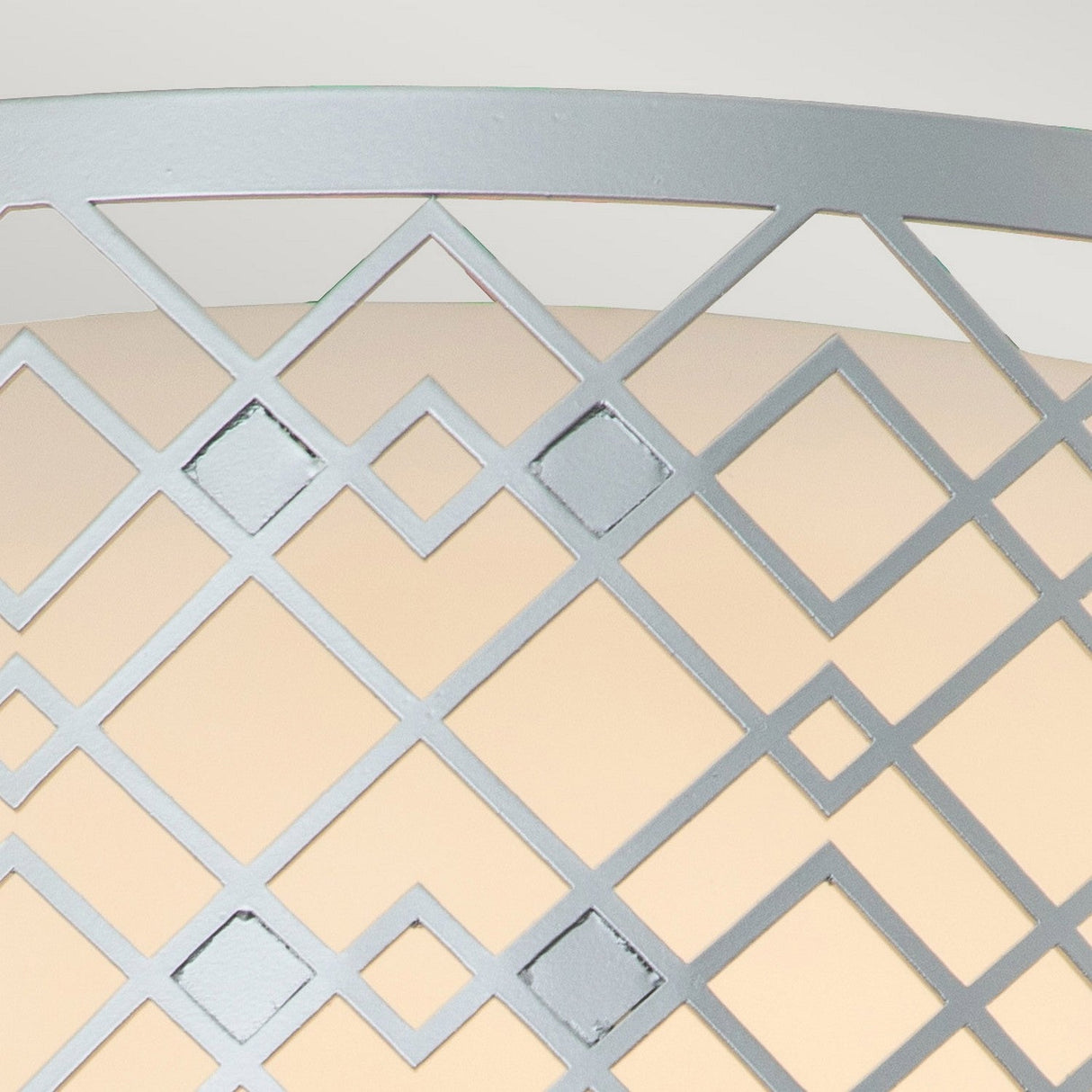 A close-up view of the Ziggy 2 Light Medium Flush showcases its geometric metal lattice pattern with interlocking diamond shapes, finished in lacquered silver. The light gray structure stands out against an off-white background, creating a striking contrast between the foreground and background elements.