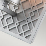Close-up of a geometric metal grate showcasing a pattern of squares and rectangles. In the top left corner, a Ziggy 1 Light Wall Light in lacquered silver catches the light, adding modern elegance to the scene. The neutral, light background further enhances its contemporary aesthetic.