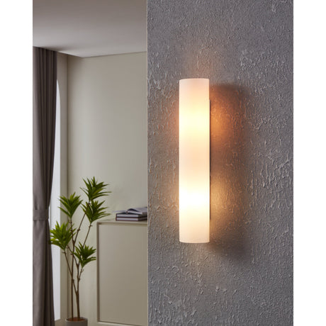 Zola Wall Light in opal glass with white steel backplate, shown lit on a textured grey wall, with a living room setting visible alongside.
